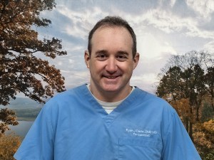 Headshot of periodontist Dr. Ryan J. Davis smiling in blue medical scrubs against an outdoor autumn landscape background